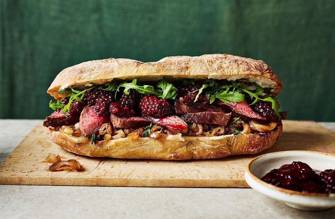 Gourmet steak sandwich on wooden board features sliced beef, blackberries, arugula, and caramelized onions with a dark green backdrop.