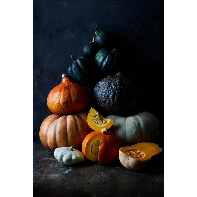 Still life photograph of various pumpkins and gourds, in shades of orange, green, and white, stacked against a dark background.