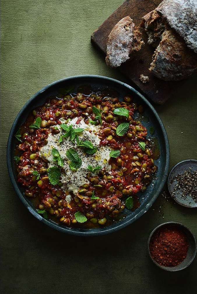 Overhead shot of a dark plate filled with a tomato and lima bean dish, topped with cream and mint, served with bread.