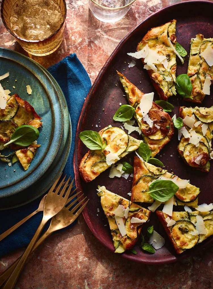 Overhead shot of zucchini frittata slices on plates and platter, garnished with basil and parmesan, on a marble surface with two forks.