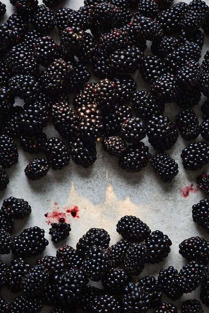 Overhead shot of many fresh blackberries scattered on a white surface, some with red juice. Food still life. Dark food photography.
