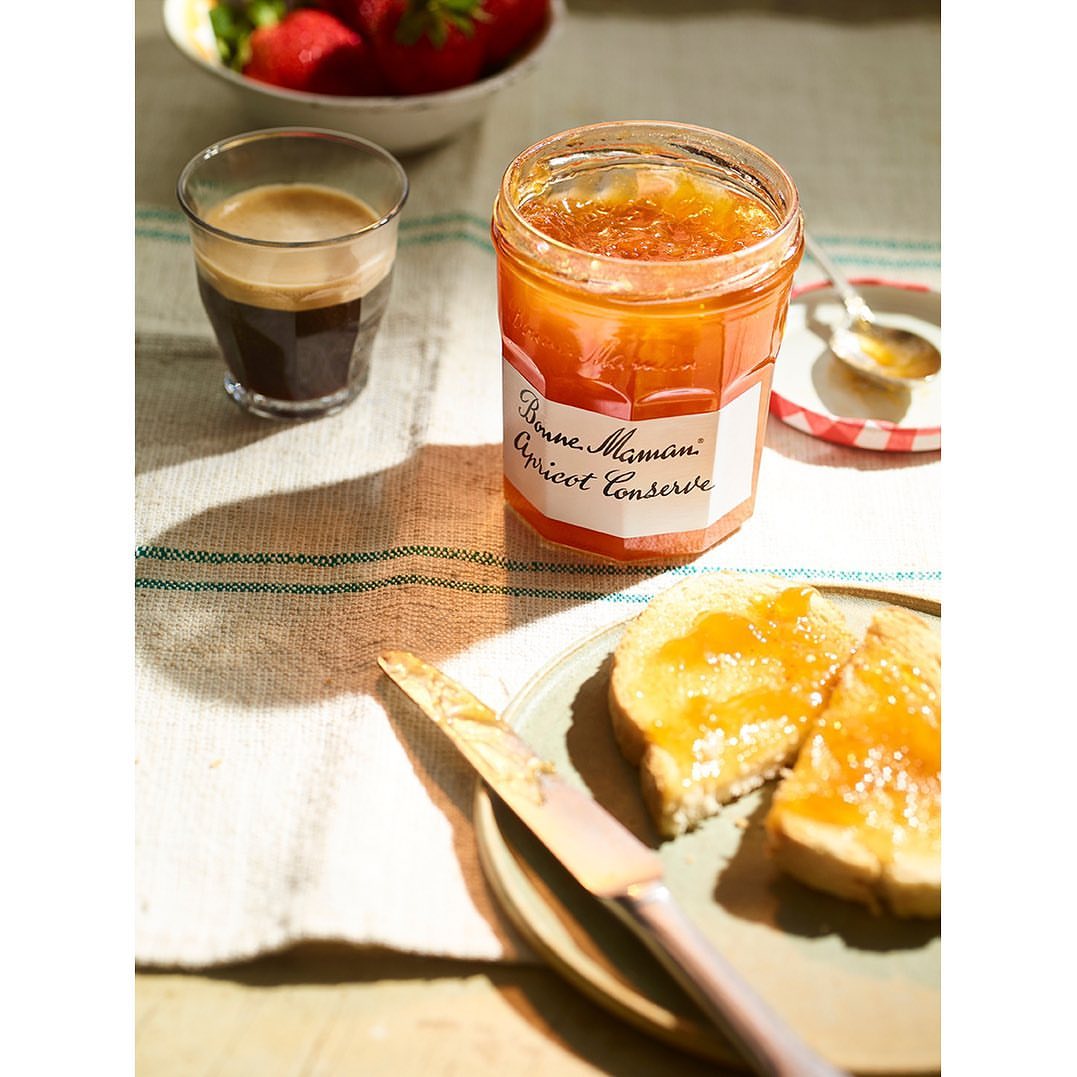 Overhead shot of apricot jam jar, espresso, strawberries, toast with jam, and knife on cloth on a wooden table, bright natural light.