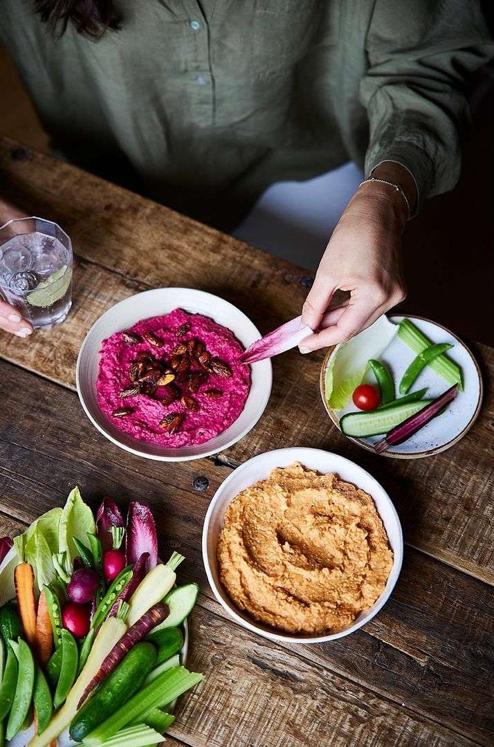 Overhead shot of a rustic wooden table with bowls of beetroot and hummus dips, surrounded by assorted raw vegetable sticks and water glass.