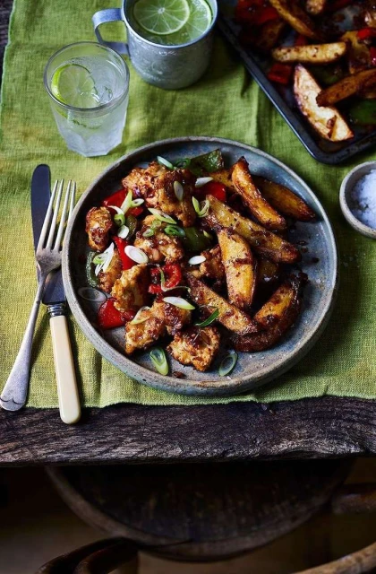 Overhead shot of chicken and potato wedges plated with drinks and cutlery on a rustic wooden surface with a green cloth.