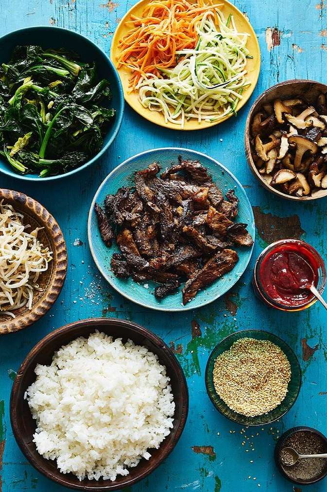 Overhead shot of bibimbap ingredients: rice, sautéed greens, meat, sprouts, julienned carrots, zucchini, mushrooms, and gochujang sauce arranged on a blue table.