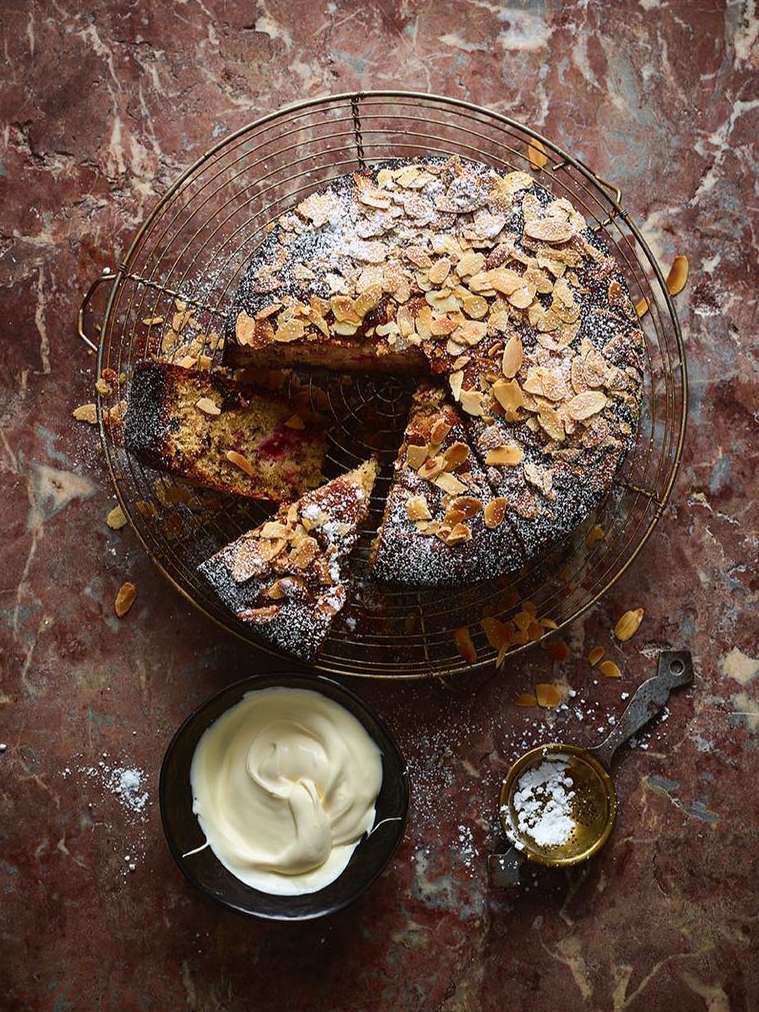 Overhead shot of almond cake on wire rack with powdered sugar, served with cream and spoon of sugar on a marble surface.
