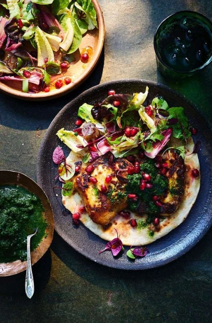Overhead shot of grilled halloumi cheese with pomegranate seeds and salad on a flatbread served on a plate with sauce and a drink.