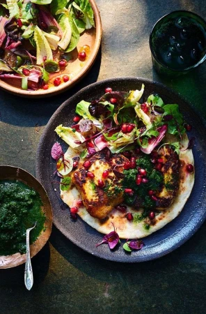 Overhead shot of grilled halloumi cheese with pomegranate seeds and salad on a flatbread served on a plate with sauce and a drink.