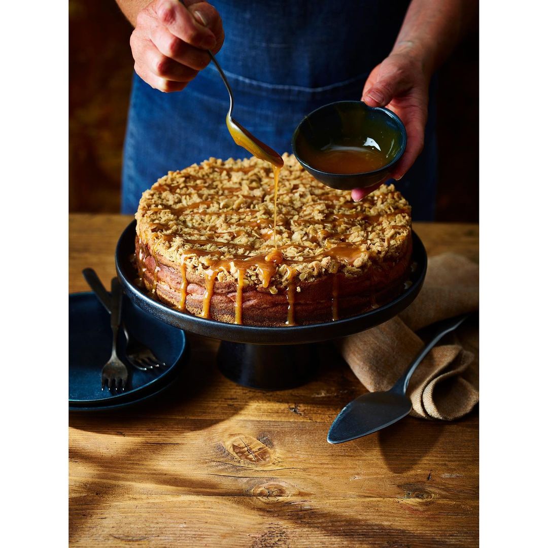 A hand pours caramel sauce over a crumble cheesecake on a black cake stand, with plates, forks, and a serving utensil on a wood surface.