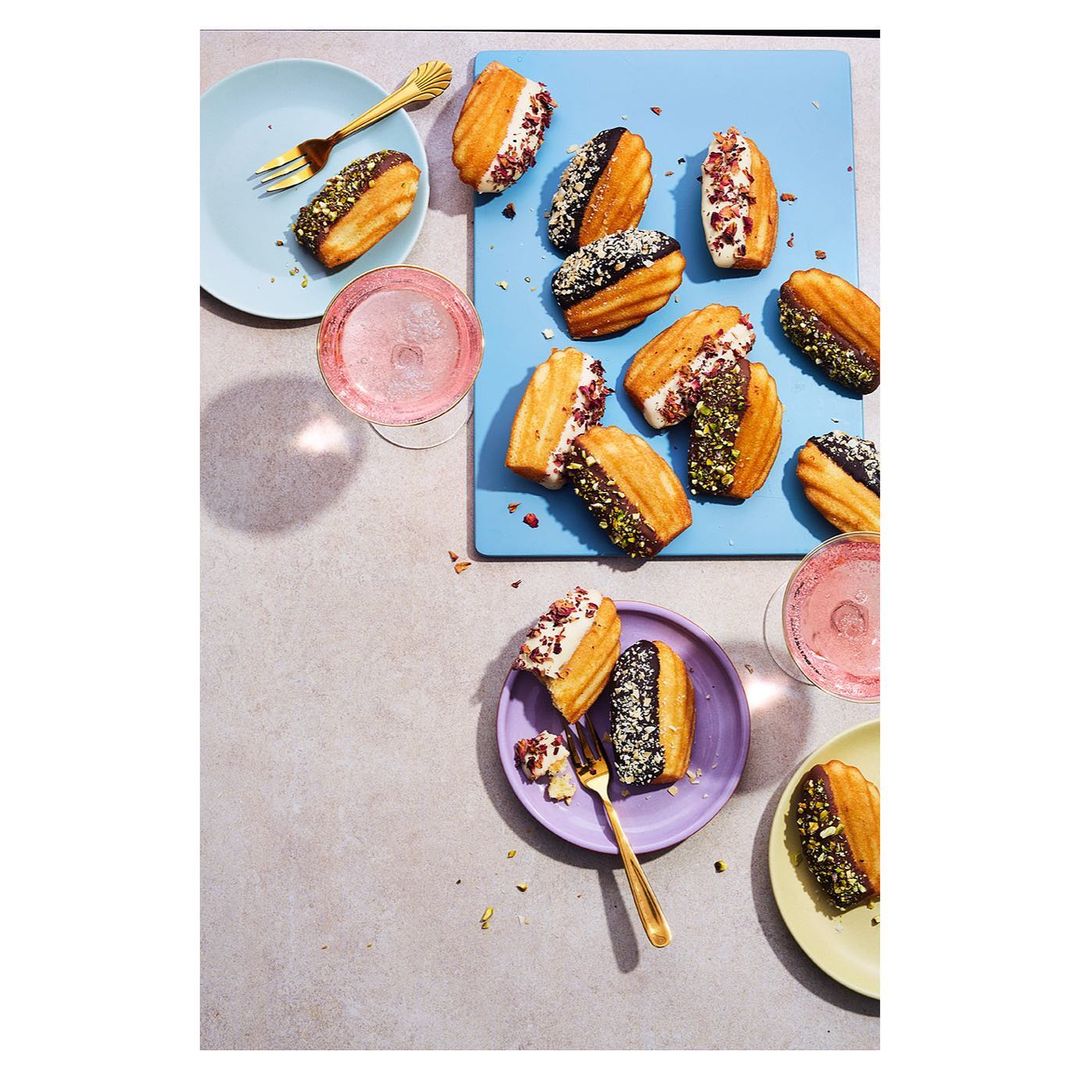 Overhead shot of colourful plates and a blue serving board with various madeleines decorated with icing and toppings next to pink cocktails.