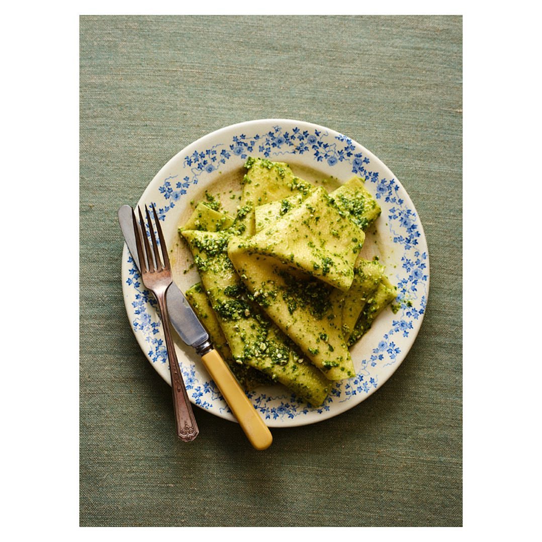 Overhead shot of plate of pasta with pesto sauce next to silverware on green tablecloth. Food photography with bright lighting.