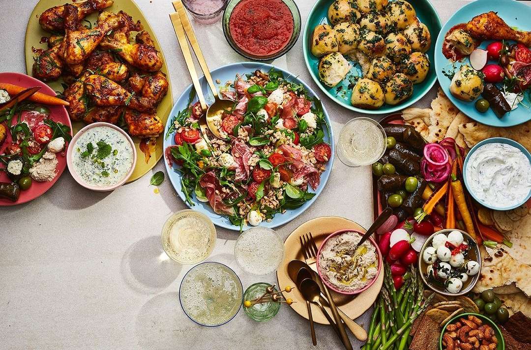 Overhead shot of a variety of vibrant foods on a table, including chicken, salad, dips, and other appetizers and snacks.
