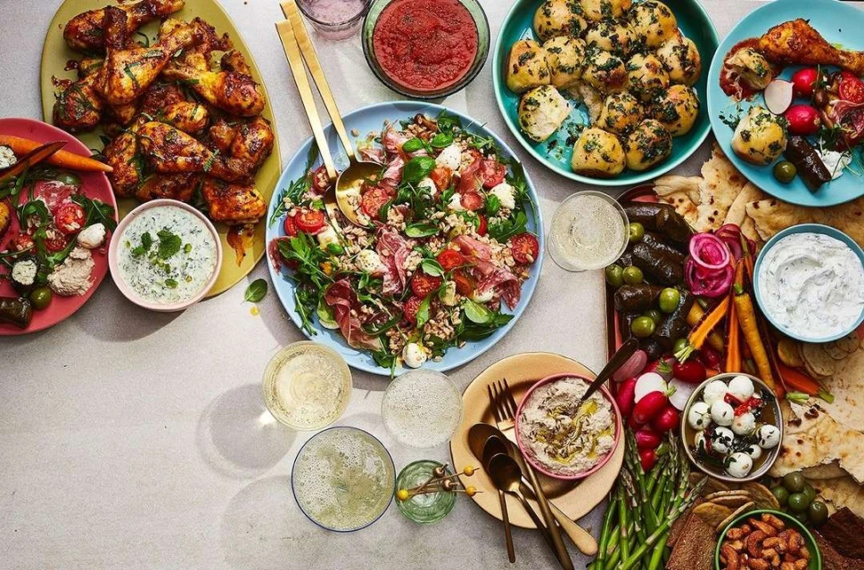 Overhead shot of a variety of vibrant foods on a table, including chicken, salad, dips, and other appetizers and snacks.