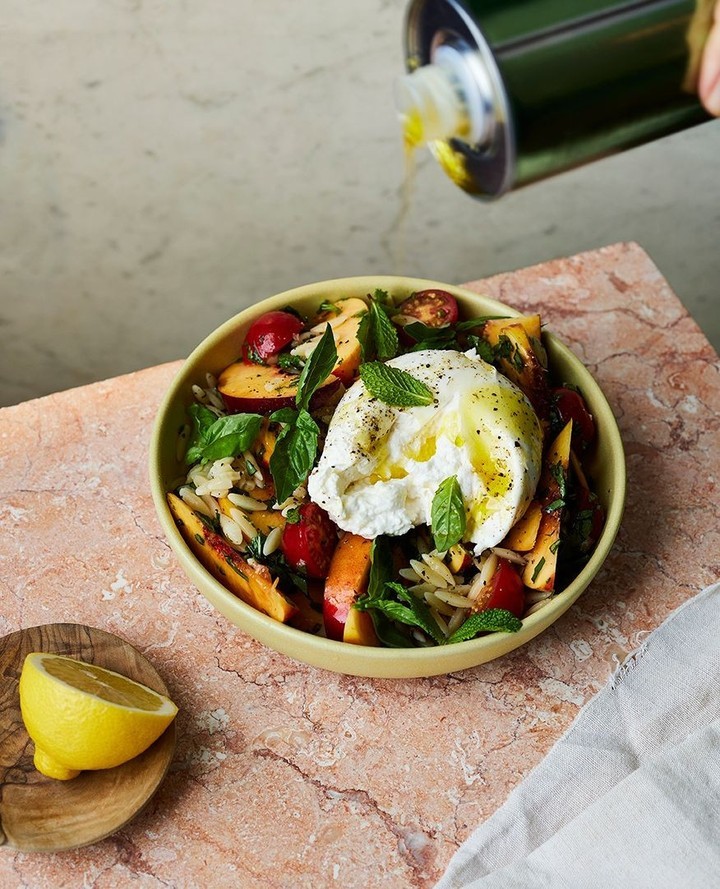 Olive oil is drizzled over a summer salad with burrata cheese, peaches, tomatoes, mint, and pasta in a yellow bowl on a marble table.