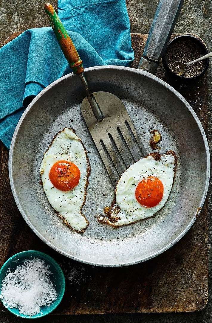 Overhead shot of two fried eggs with bright orange yolks in a skillet with a spatula, salt, pepper, and a blue cloth.
