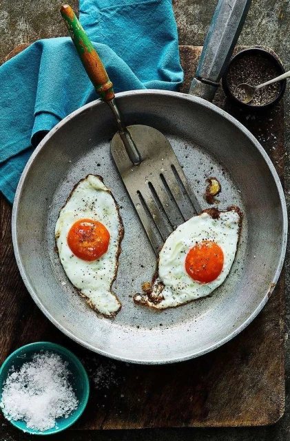 Overhead shot of two fried eggs with bright orange yolks in a skillet with a spatula, salt, pepper, and a blue cloth.