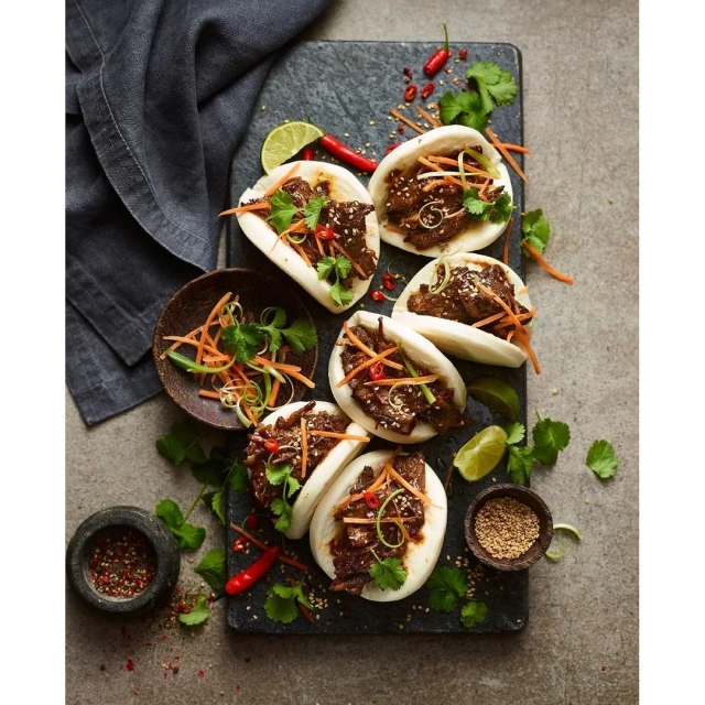 Overhead shot of a variety of bao buns filled with meat, garnishes, and sauces, arranged on a gray stone platter with limes and spices.