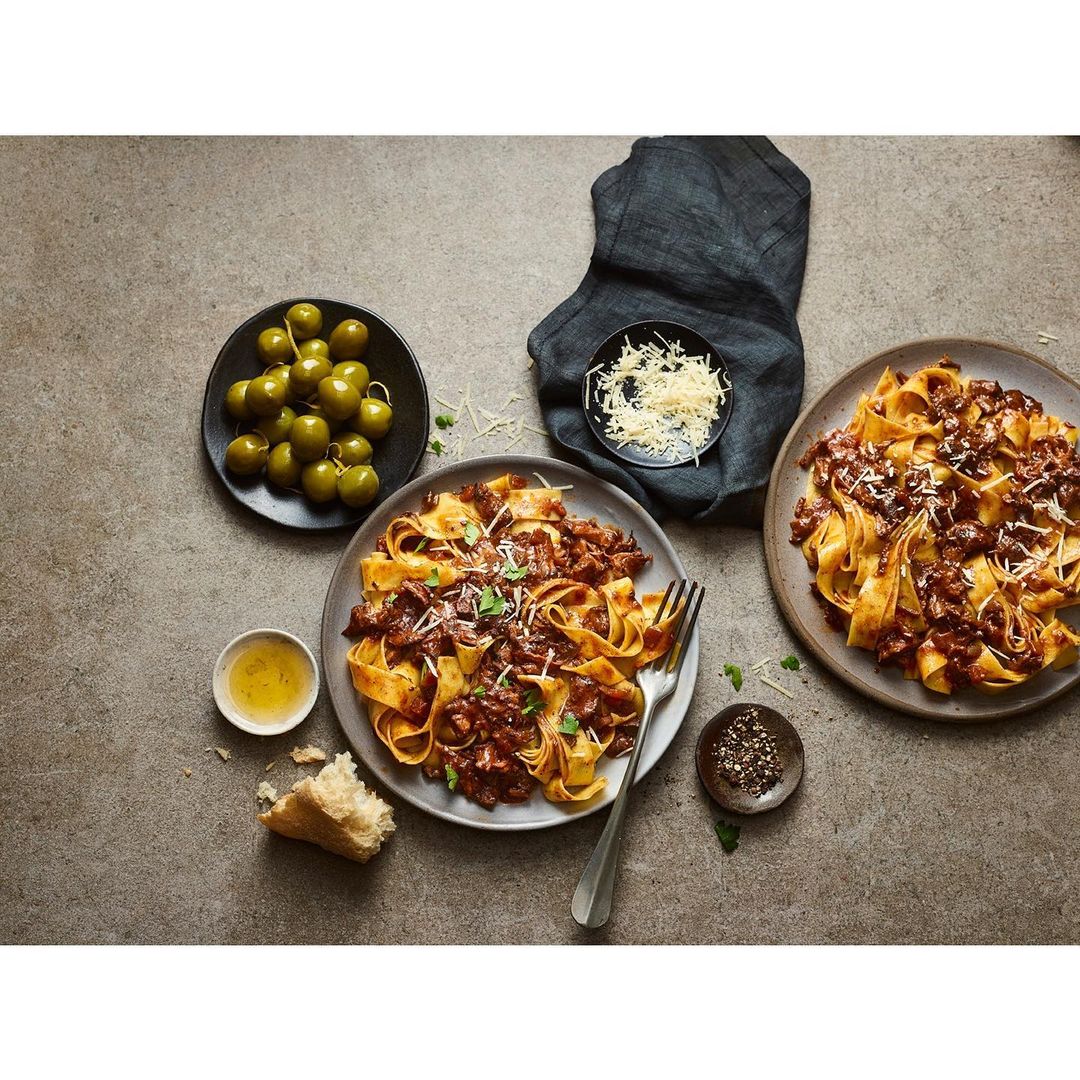 Overhead shot of two plates of pasta with meat sauce, bowl of olives, cheese, bread, pepper, and olive oil on grey surface.
