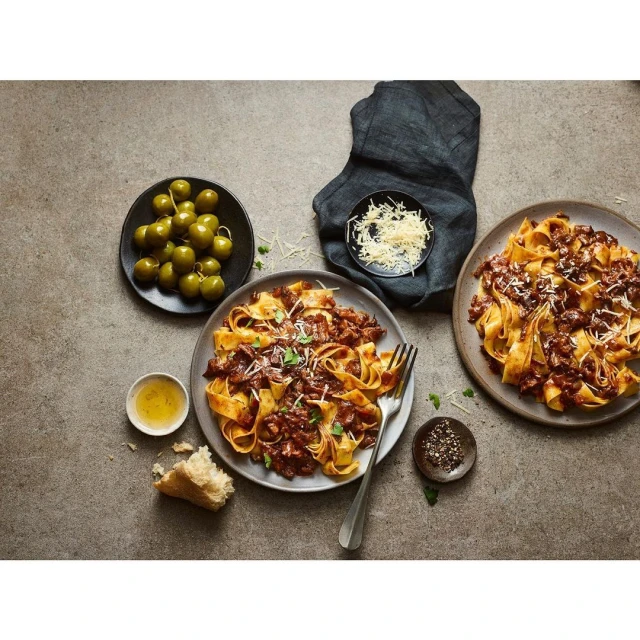 Overhead shot of two plates of pasta with meat sauce, bowl of olives, cheese, bread, pepper, and olive oil on grey surface.