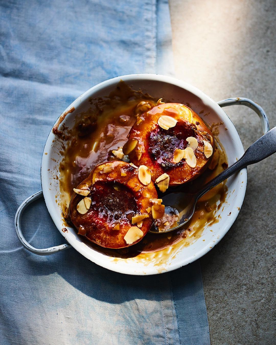 Overhead shot of two peach halves baked with almonds in a white dish, glazed with syrup, served with a silver spoon on a blue tablecloth.