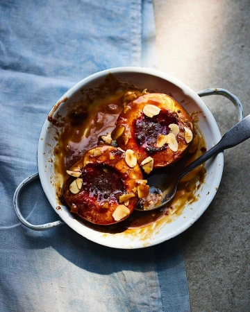 Overhead shot of two peach halves baked with almonds in a white dish, glazed with syrup, served with a silver spoon on a blue tablecloth.