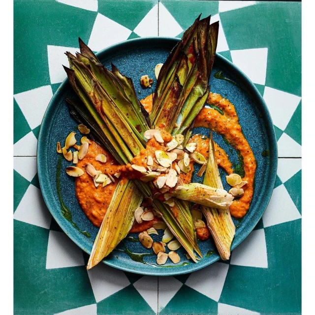 Overhead shot of leeks with sauce and almonds on a blue plate, set on a green and white patterned tile surface. Food photography.