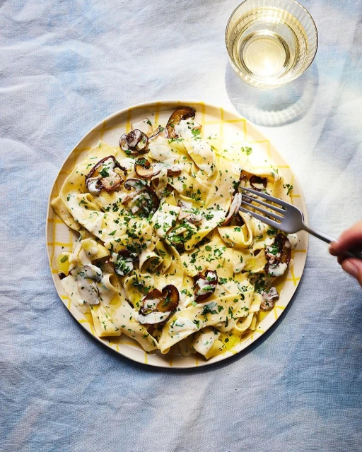Overhead shot of a plate of mushroom pappardelle pasta with fresh parsley on a blue tablecloth next to a glass of white wine.