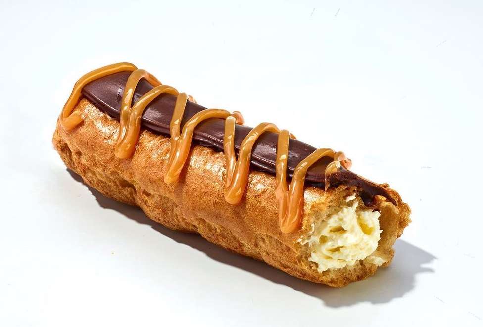 Close-up of an éclair pastry filled with cream and topped with chocolate ganache and caramel drizzle on a white background.