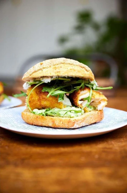 Delicious fish burger on a plate featuring a crispy fish fillet, fresh arugula, and a creamy sauce on a wooden table in an indoor setting.