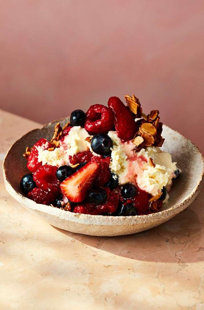 Close-up of mixed berries, cream, and almonds in a beige ceramic bowl against a pink backdrop, lit by soft, diffused lighting.