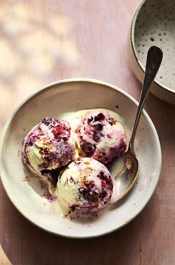 Three scoops of berry ice cream in a ceramic bowl with a spoon, sitting on a light pink surface with soft shadows.
