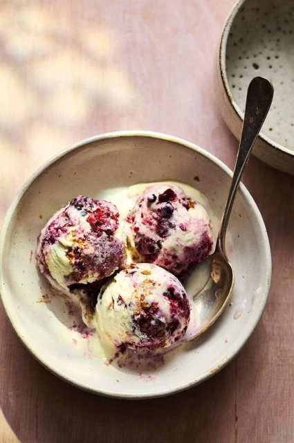 Three scoops of berry ice cream in a ceramic bowl with a spoon, sitting on a light pink surface with soft shadows.
