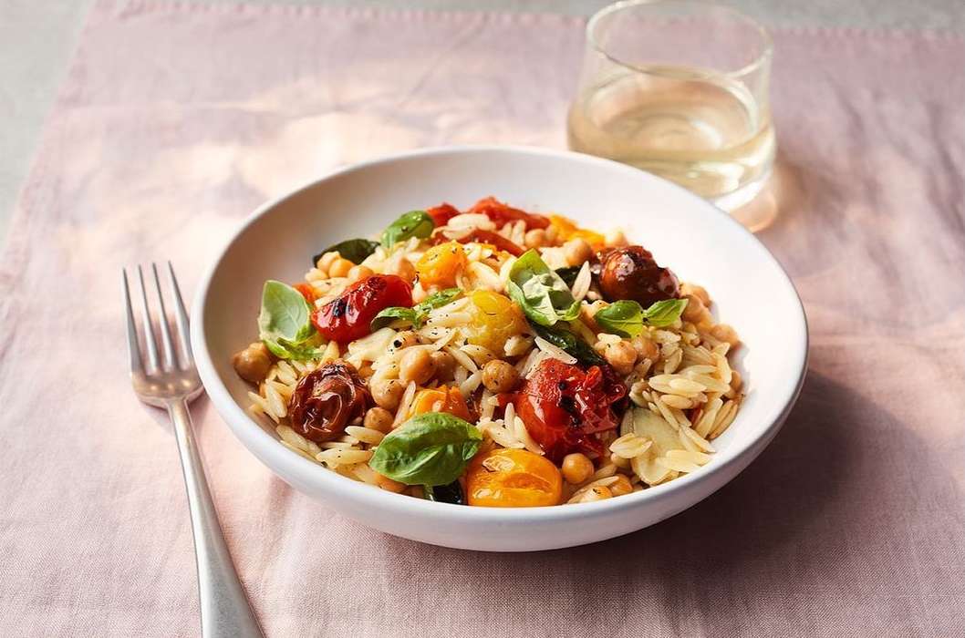Overhead view of orzo pasta salad with tomatoes, chickpeas, basil, and shaved cheese in a white bowl with a fork and drink.