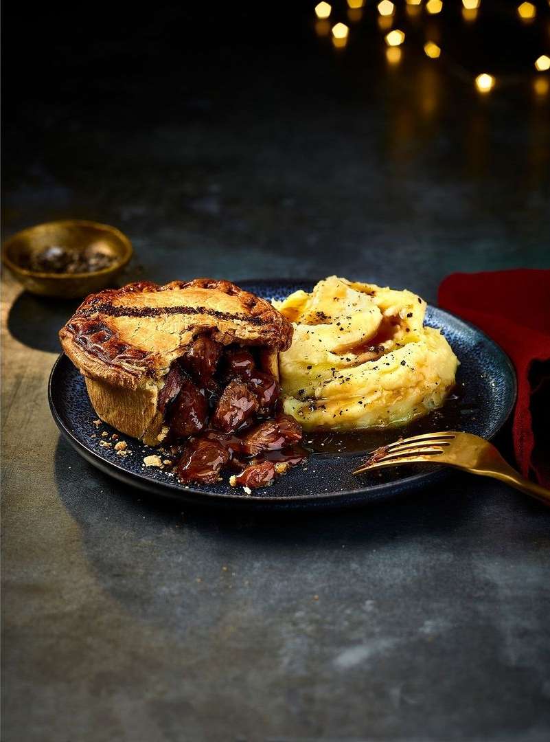 Close-up of a savory meat pie with flaky crust alongside mashed potatoes on a dark plate, with a gold fork and blurred lights.