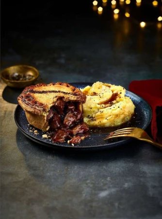 Close-up of a savory meat pie with flaky crust alongside mashed potatoes on a dark plate, with a gold fork and blurred lights.