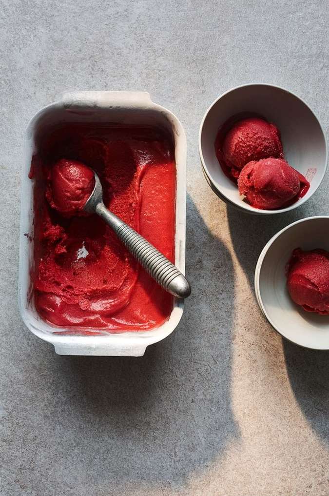 Overhead shot of red sorbet in a white container, scooped with a metal spoon, and two bowls of sorbet on a gray surface.