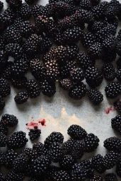 Close-up overhead view of fresh dark blackberries scattered on a light grey stone surface with scattered juice stains.