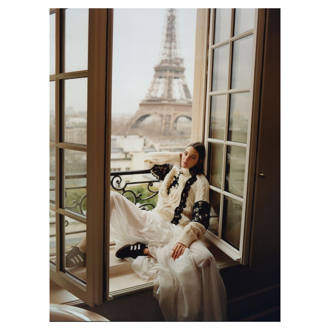 Fashion shot of a woman in a dress and sweater sitting in a window with the Eiffel Tower in the background on a cloudy day.