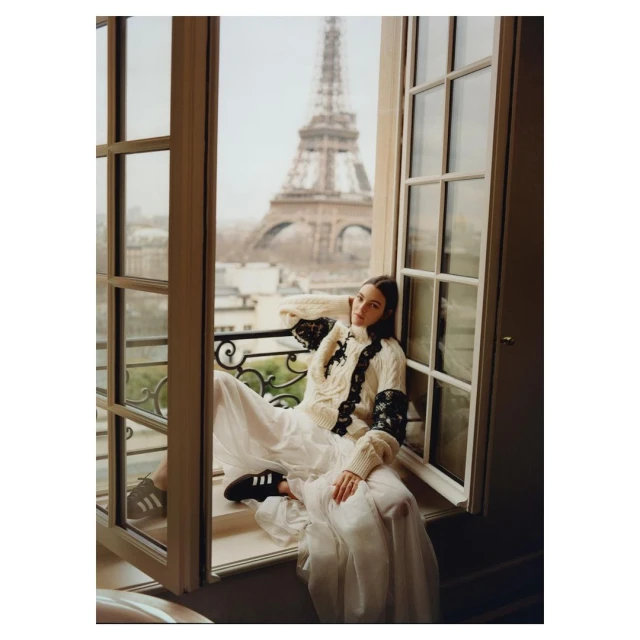 Fashion shot of a woman in a dress and sweater sitting in a window with the Eiffel Tower in the background on a cloudy day.