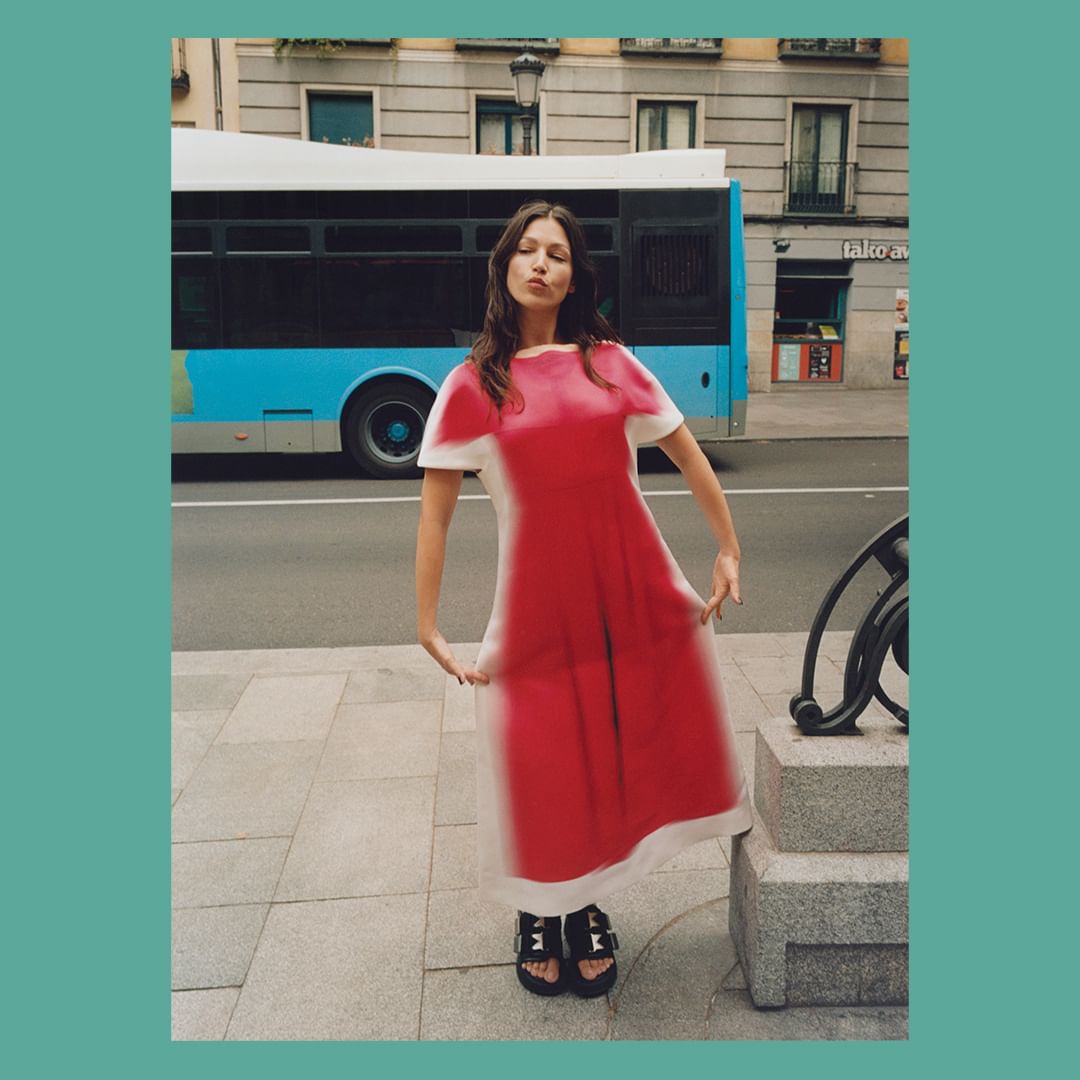 Adult woman in a bright red and white dress poses in an urban setting with a city bus passing by on a sunny day.