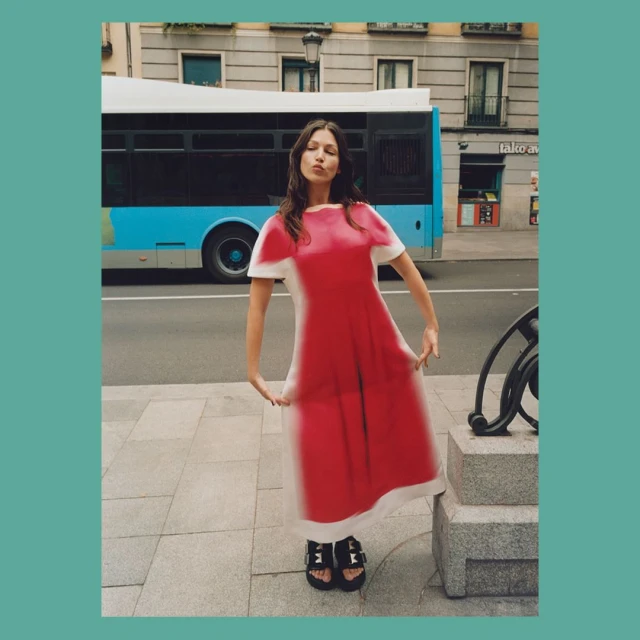 Adult woman in a bright red and white dress poses in an urban setting with a city bus passing by on a sunny day. 