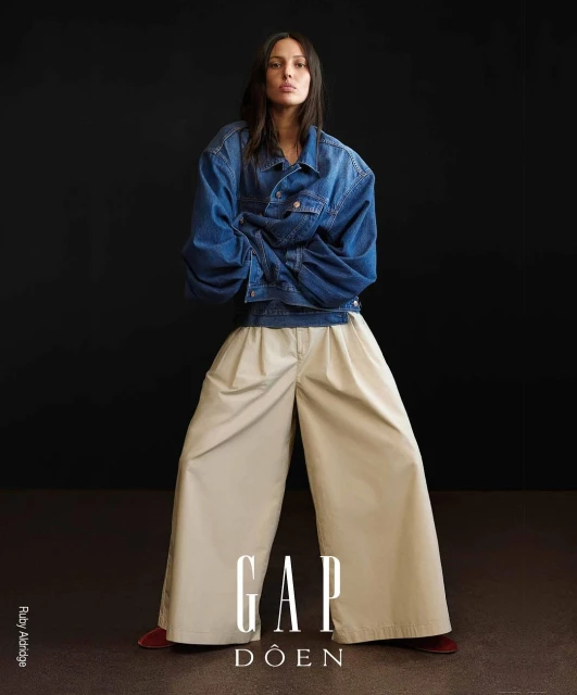 Fashion editorial shot: a stylish woman in a denim jacket and wide-leg trousers strikes a pose against a dark backdrop.