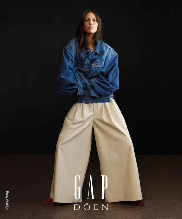Fashion editorial shot: a stylish woman in a denim jacket and wide-leg trousers strikes a pose against a dark backdrop.
