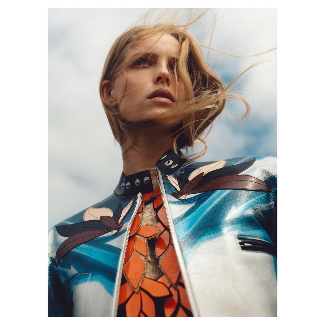 Woman with fair hair wearing a uniquely patterned blue, white, and red leather jacket looking into the distance against a cloudy sky.