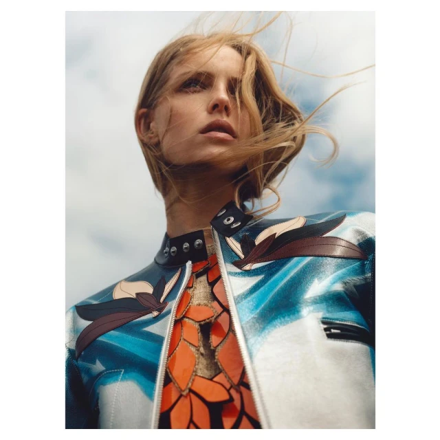 Woman with fair hair wearing a uniquely patterned blue, white, and red leather jacket looking into the distance against a cloudy sky.