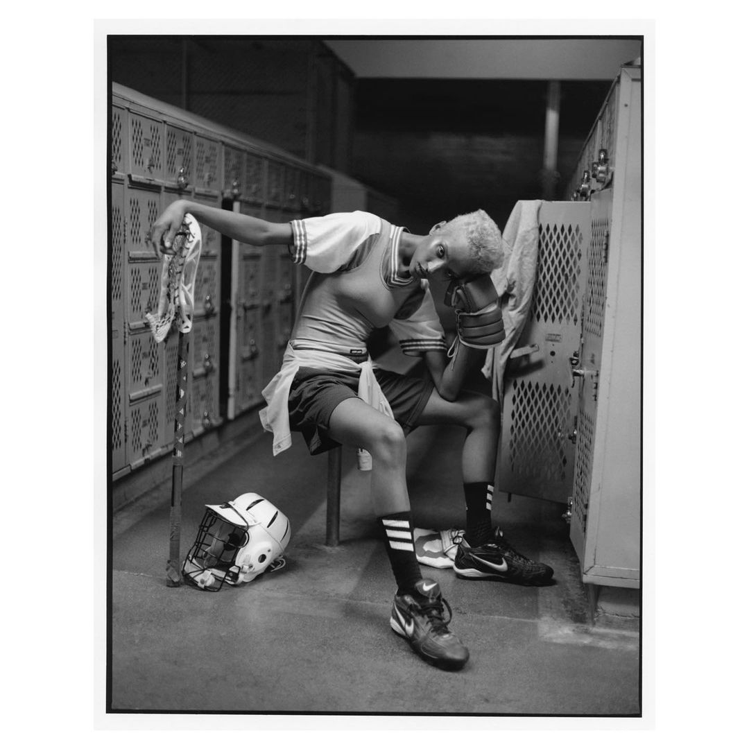 Black and white portrait of a tired lacrosse player sitting in the locker room, holding a lacrosse stick, with equipment and lockers in view.