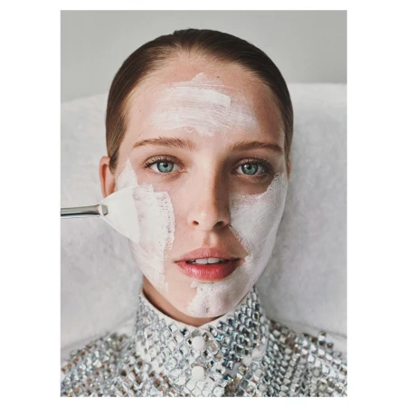 Close-up of a woman with white facial mask applied with spatula in a beauty salon setting; she wears a silver embellished top.