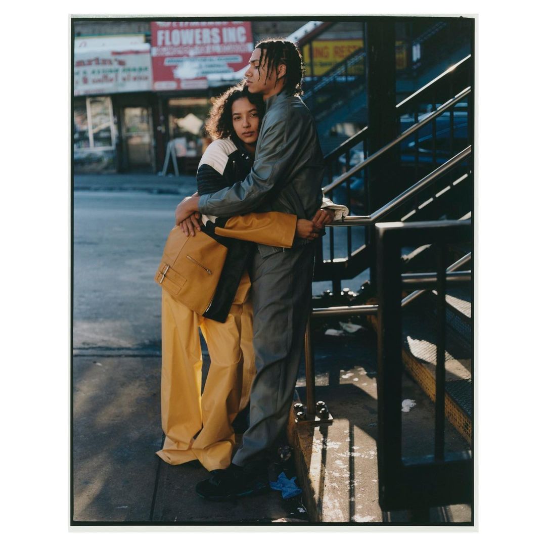 Couple embracing on a city street with a brick building and metal stairway in the background, wearing jackets and pants.