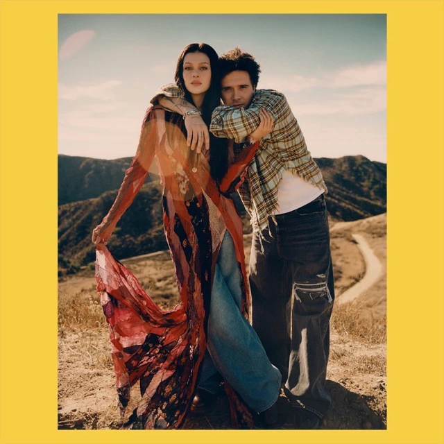 Couple poses outdoors in fashion shot on hillside. Woman wears sheer gown, man in flannel. Hilly background, portrait format.