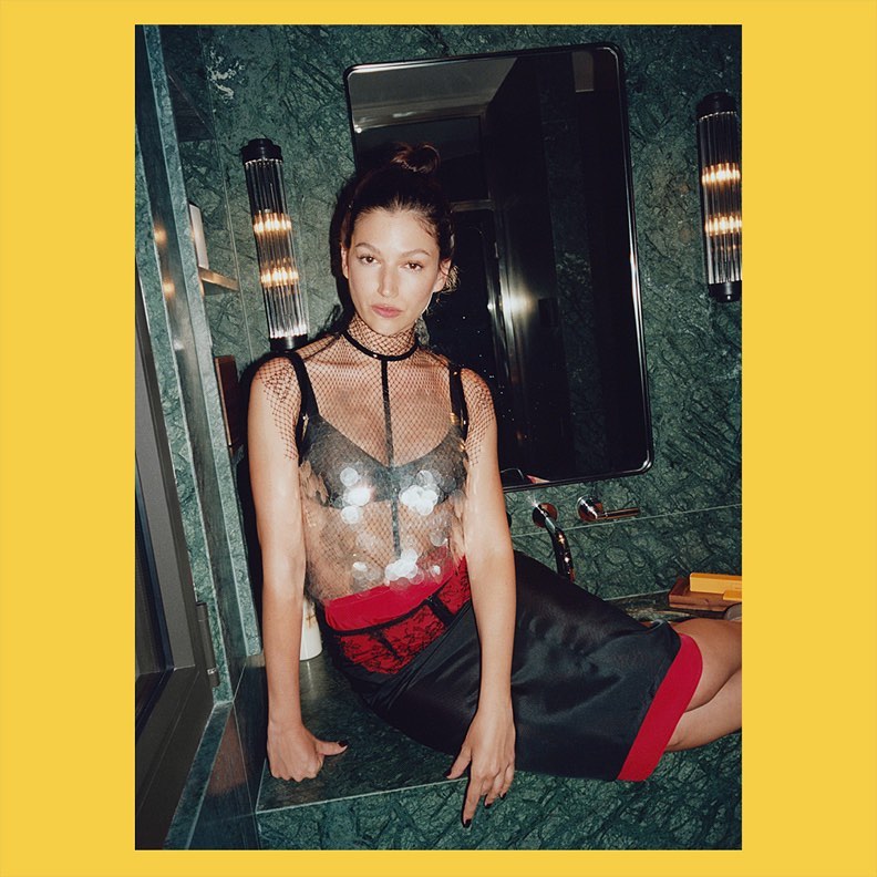 Adult female model in a mesh top and black skirt sitting on a green marble counter in a bathroom with sconces and a mirror.